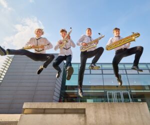 Junge Musiker:innen springen vor einem Gebäude mit ihren Instrumenten von einer Mauer